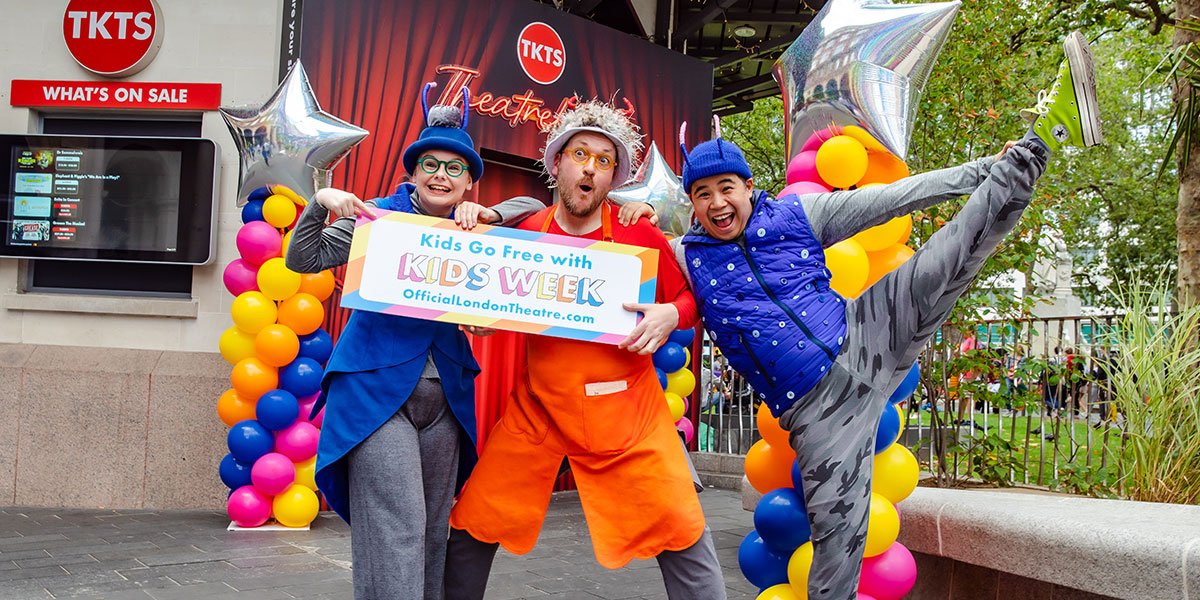 Three people stand, one in the middle wearing orange and one either side wearing blue. The person on the right has kicked their leg up in the air