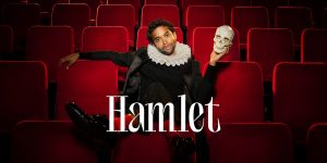 The image is set in a theatre with red velvet seats. A man sits casually across the theatre chairs, dressed in a modern black outfit with a traditional Elizabethan ruff. He strikes a playful yet thoughtful pose, holding a skull in his left hand. His expression is poised, with a slight smirk. The title “Hamlet” is prominently displayed in elegant white typography near the bottom center.