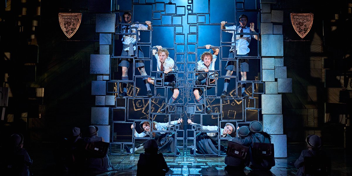 Children in school uniforms climb a geometric metal structure on stage, illuminated by blue lighting.