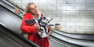 A person multitasking on an escalator, holding a small dog, a tablet, and multiple coffee cups, dressed in a red coat, with a tube station backdrop.