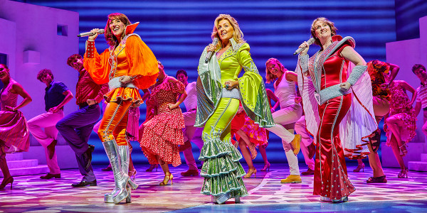 Kate Graham as Tanya, Mazz Murray as Donna, Nicola-Dawn Brook as Rosie and Company in MAMMA MIA! Kate, Mazz and Nicola-Dawn are each wearing an orange, green and red flared jumpsuit. They are singing and the Company is dancing behind them.